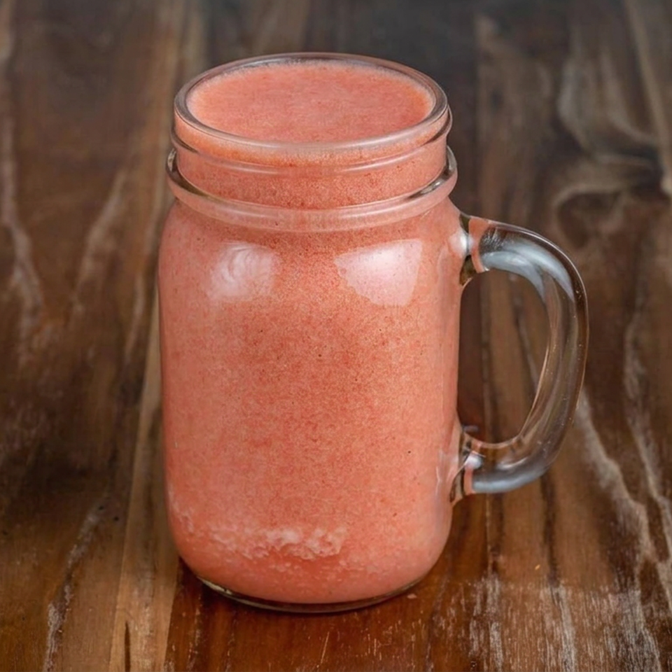 Pink smoothie in a clear handled glass jar on a rustic wooden table.